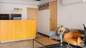 Zona de recepción con un separador amarillo, mostrador de recepción, área de descanso con una silla azul y un sofá naranja, y un reloj fijado en la pared.