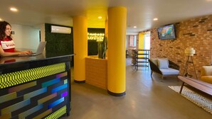 A reception desk with a colorful, geometric design. There is a woman using a laptop at the desk. The lobby features yellow pillars, a brick wall, and a seating area with a sofa and chairs. - Hotel Estancias VIVE MX Cuauhtemoc, Trademark by Wyndham (Mexico City)