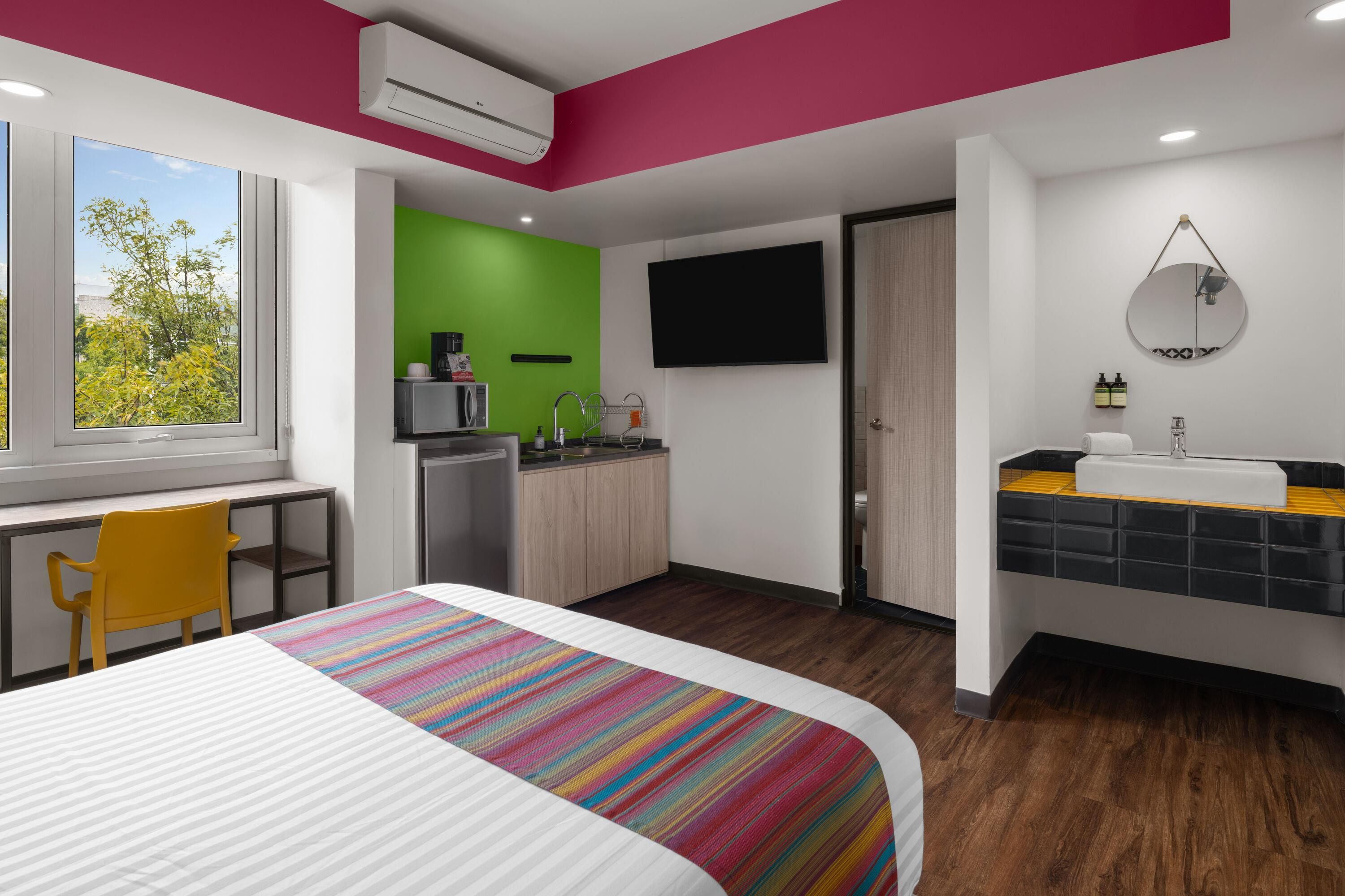 Quarto de hotel moderno com cama, escrivaninha, cozinha compacta e banheiro.
