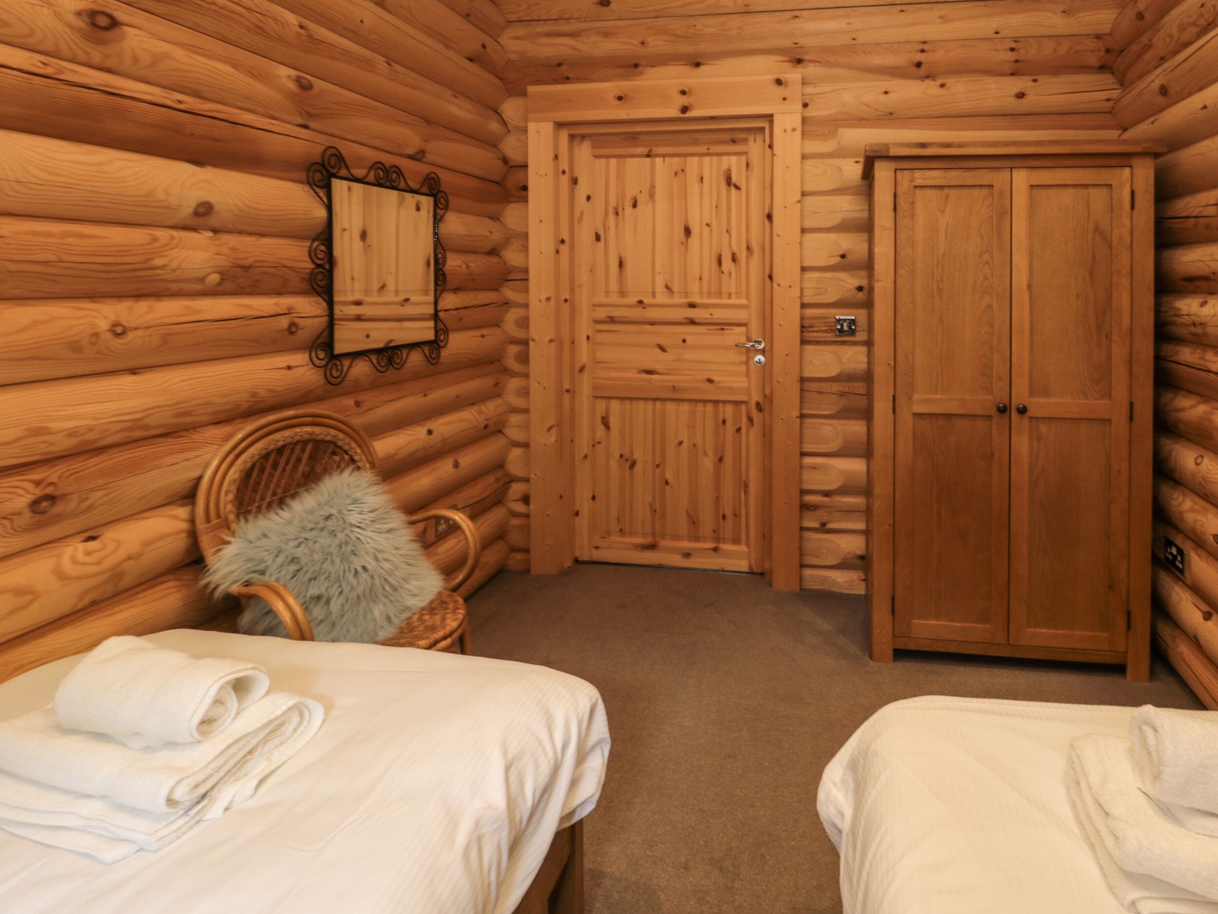A log cabin room with two beds, a wooden door, a mirror, and a wicker chair with a fur throw.