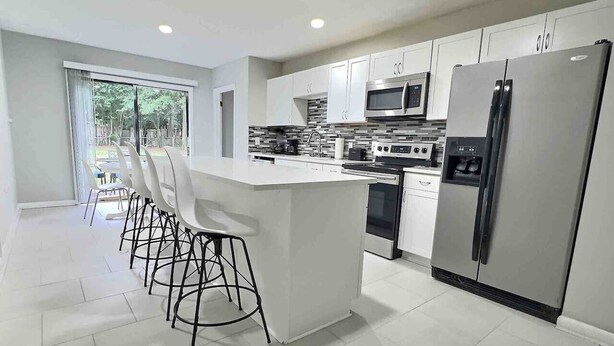 A modern kitchen with white cabinets, a central island with bar stools, stainless steel appliances, and a large window providing natural light.