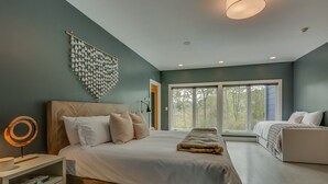 A bedroom with a large bed, a bedside table, a wall-mounted light fixture, and a window with a view of trees. - Modern Country Home with Pool, Hot Tubs & Fire Pit - close to wineries & beach (Berrien Springs)