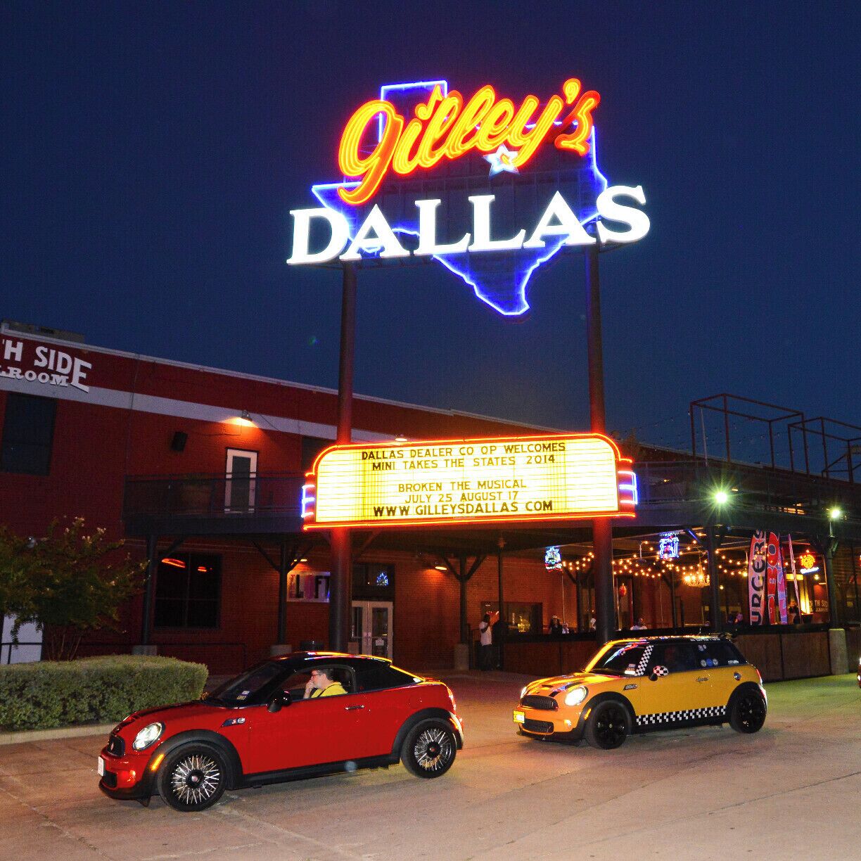 Um letreiro de neon para 'Gilley's Dallas' com uma marquise anunciando 'Dallas Dealer Co Op Welcomes Mini Takes the States 2014'. Dois carros estão estacionados na frente.