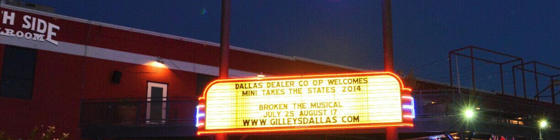 Um letreiro de neon para 'Gilley's Dallas' com uma marquise anunciando 'Dallas Dealer Co Op Welcomes Mini Takes the States 2014'. Dois carros estão estacionados na frente.