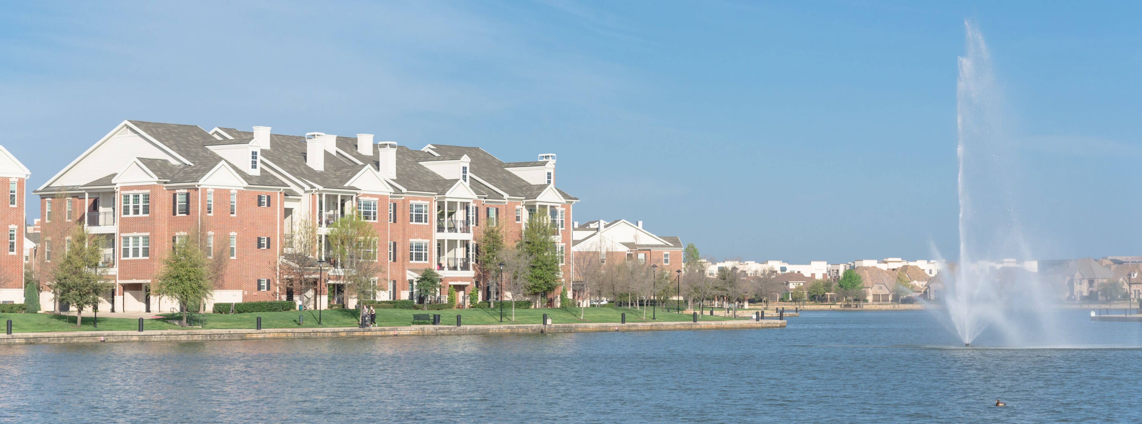 A waterfront with a row of brick buildings, a fountain, and a clear sky.