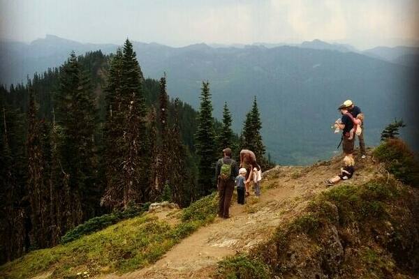 A group of people hiking on a mountain trail with dense forests and distant mountains in the background.