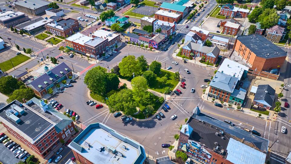 An aerial view of a city intersection with surrounding buildings, roads, and a central green space.
