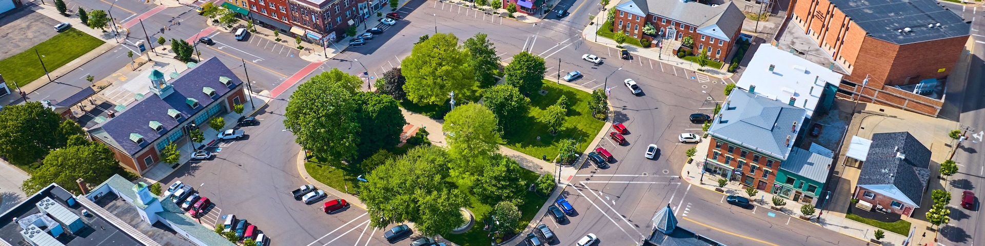 An aerial view of a city intersection with surrounding buildings, roads, and a central green space.