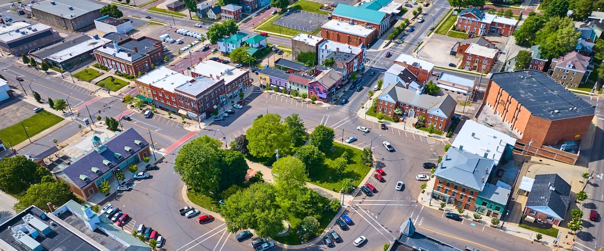 An aerial view of a city intersection with surrounding buildings, roads, and a central green space.