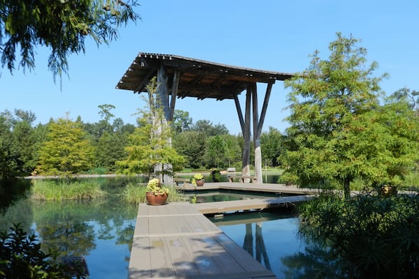 A wooden deck over a pond with a gazebo, surrounded by lush greenery and trees.