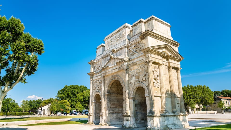 An ancient stone archway with intricate carvings and columns, surrounded by trees and a clear blue sky.