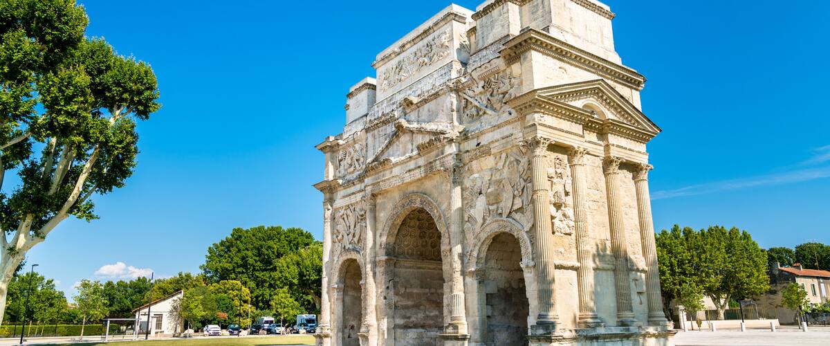 An ancient stone archway with intricate carvings and columns, surrounded by trees and a clear blue sky.