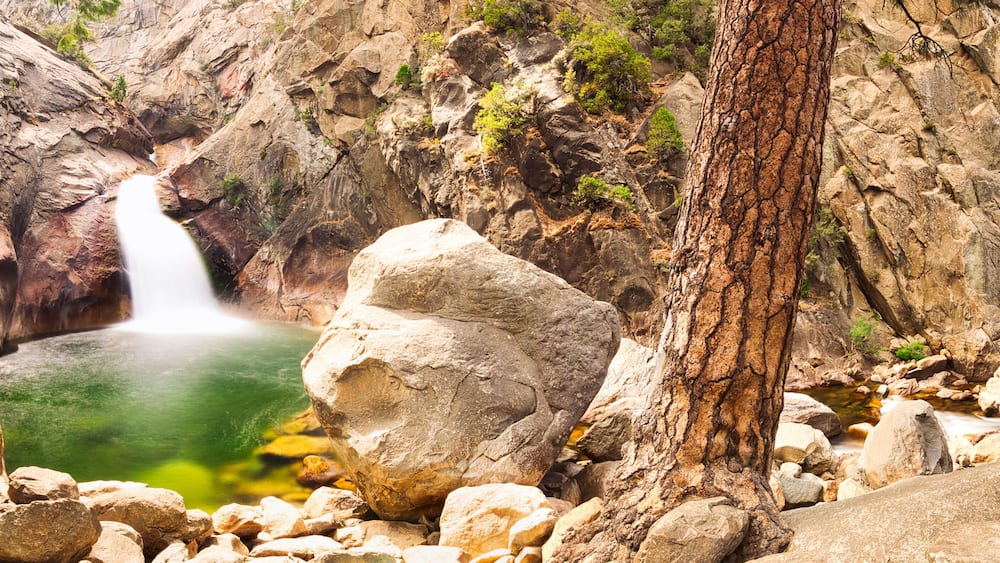 A waterfall cascading into a pool surrounded by rocky terrain and sparse vegetation.