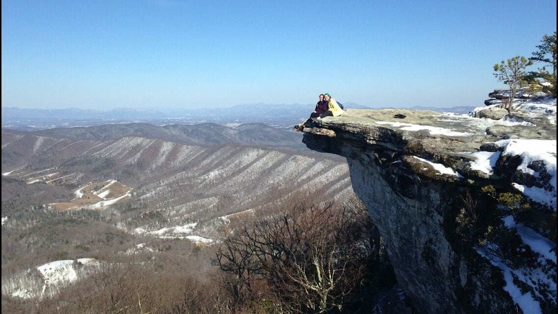 A rocky cliff edge with a person sitting on it, overlooking a valley with snow-covered hills.