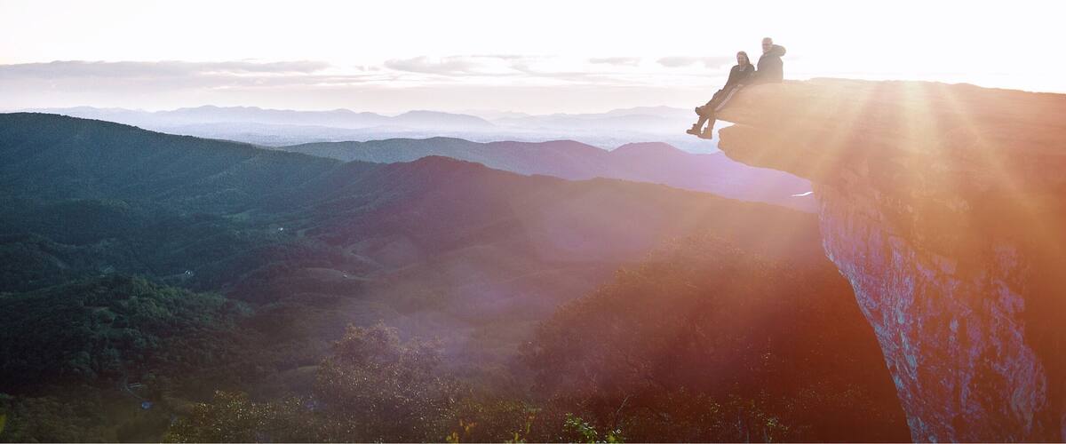 A couple sitting on a cliff edge with a view of distant mountains and a sunrise.