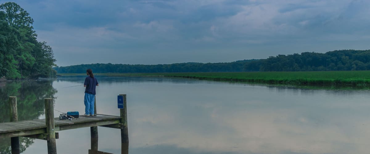 A calm lake with a wooden dock, a person fishing, and a forested hill in the background.