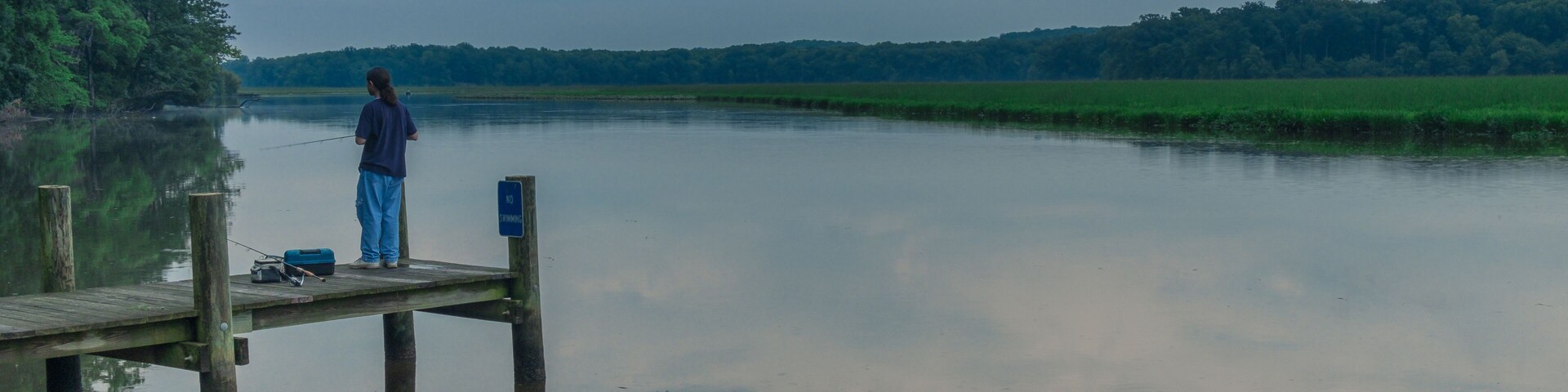 A calm lake with a wooden dock, a person fishing, and a forested hill in the background.