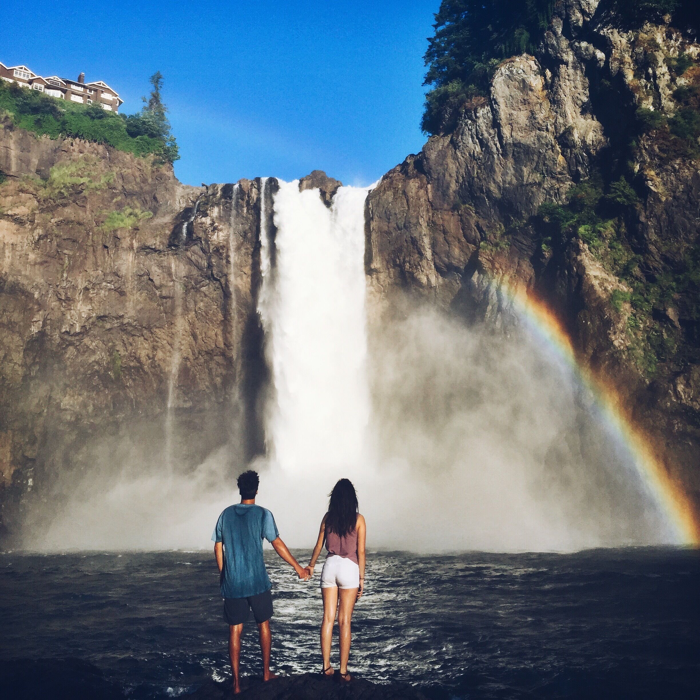 A couple holding hands in front of a waterfall with a rainbow.