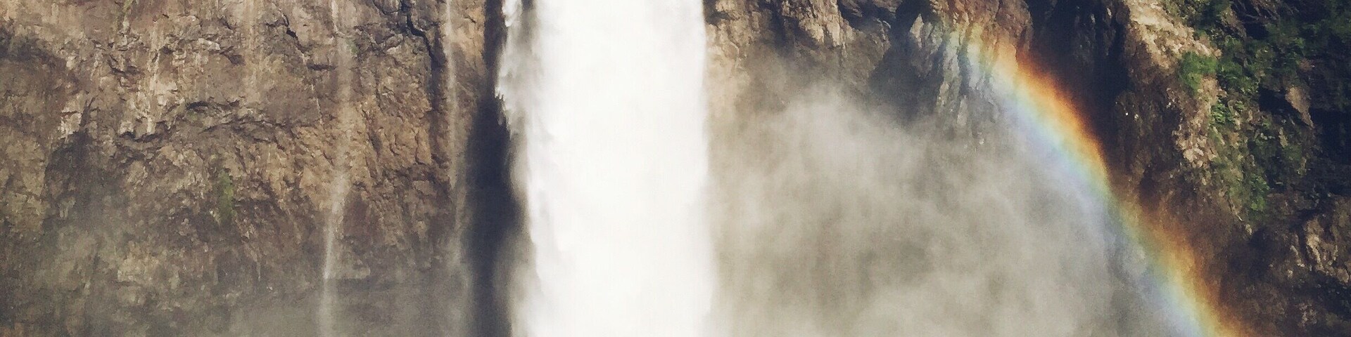 Um casal de mãos dadas em frente a uma cachoeira com arco-íris.