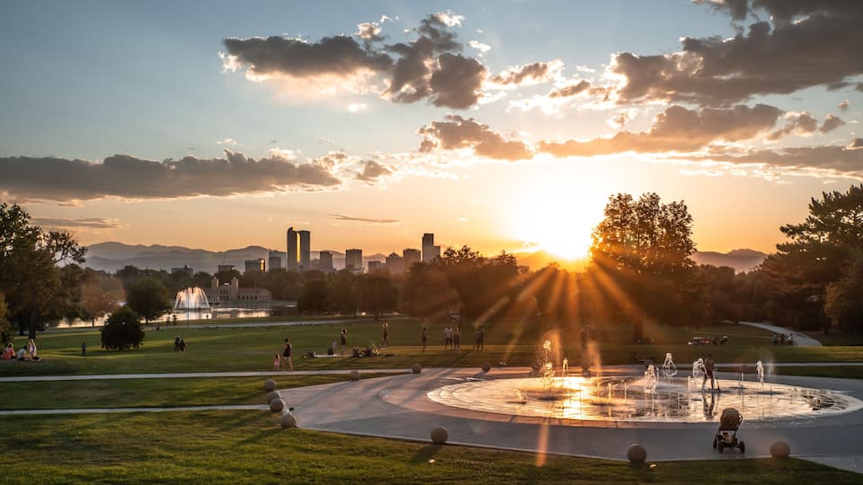 A park with a fountain, grassy areas, and a city skyline in the background during sunset.