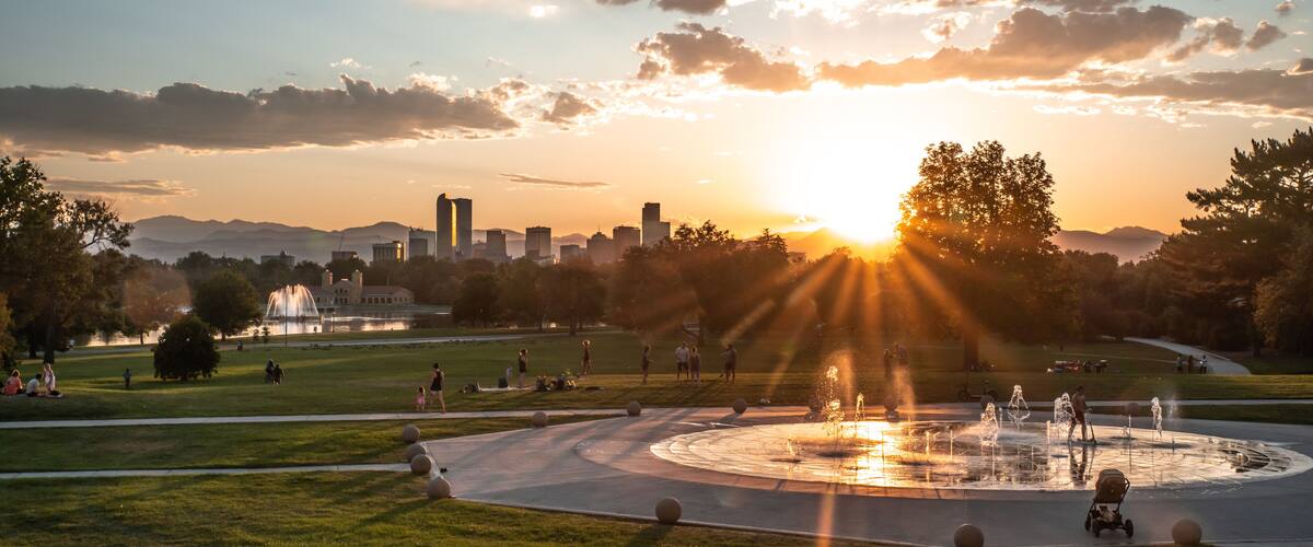 A park with a fountain, grassy areas, and a city skyline in the background during sunset.