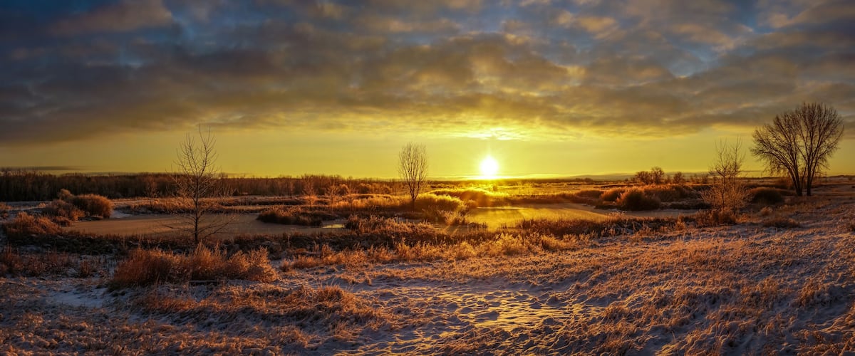 A sunset over a snowy landscape with a river and sparse trees.