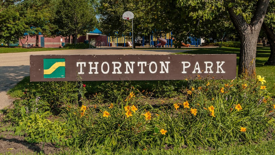 A sign for Thornton Park with a basketball hoop and playground in the background.