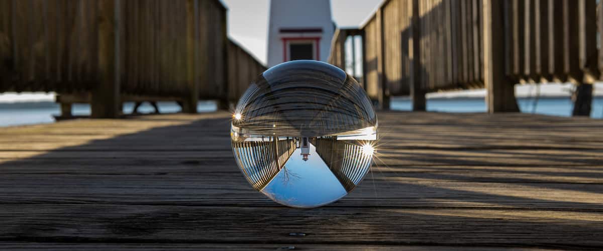 Una pasarela de madera con una esfera reflectante en primer plano y un faro al fondo.