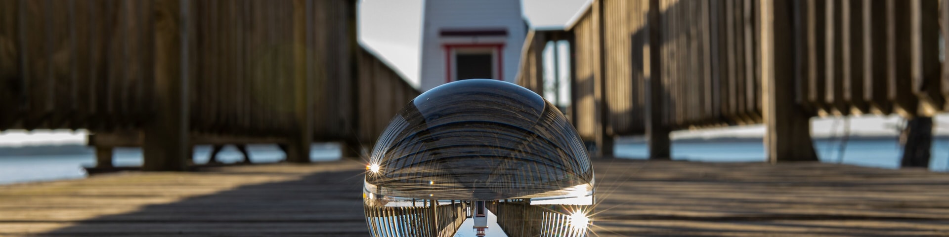 Um calçadão de madeira com uma esfera refletora em primeiro plano e um farol ao fundo.