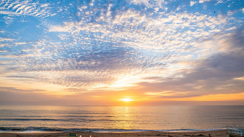 A beach with a wooden walkway leading to the water, a sunset, and a clear sky with clouds.