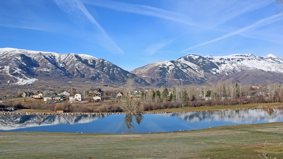 A lake reflecting snow-capped mountains, a small town with houses, and a clear blue sky.