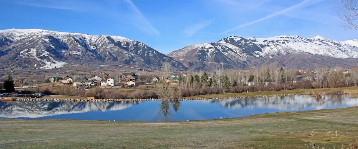 A lake reflecting snow-capped mountains, a small town with houses, and a clear blue sky.