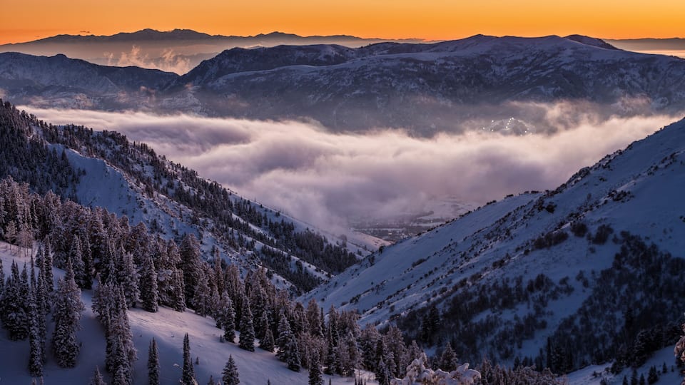A snow-covered mountain range at sunset with a valley of clouds below.