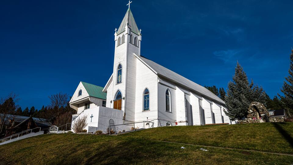 A white church with a green roof and a tall steeple on a grassy hill.