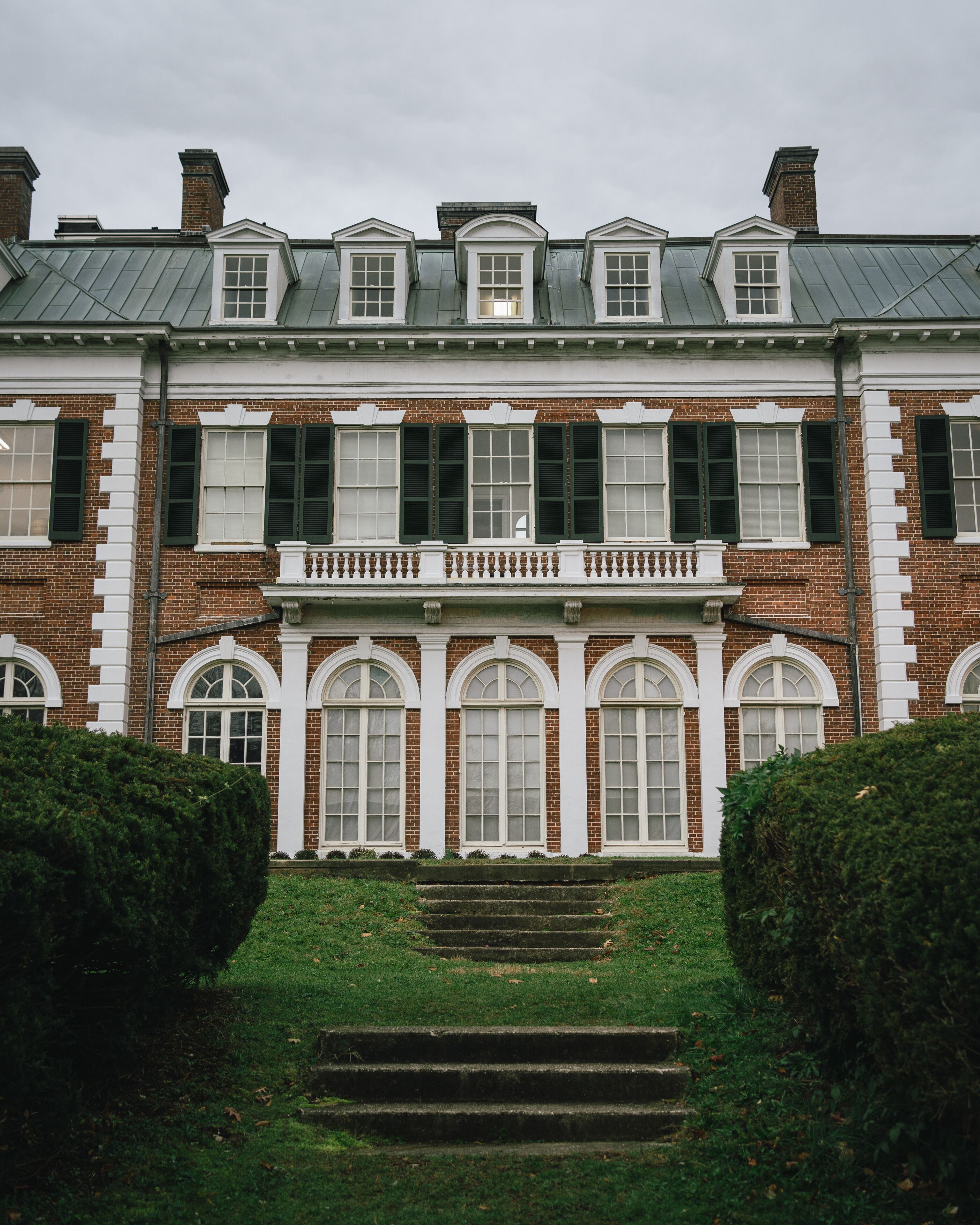 A large brick building with multiple windows, a central balcony, and a well-maintained lawn with steps leading to the entrance.