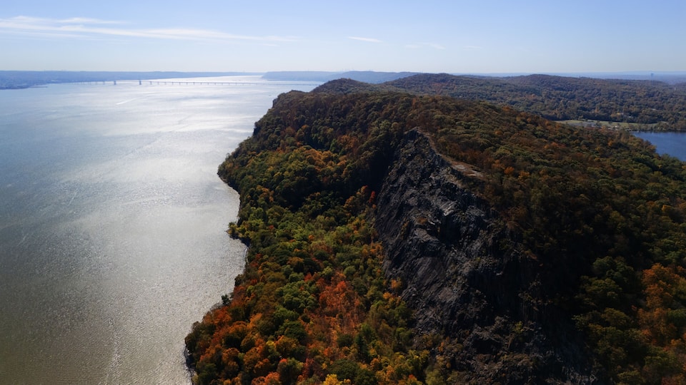 A river bordered by a forested cliff and a distant bridge.