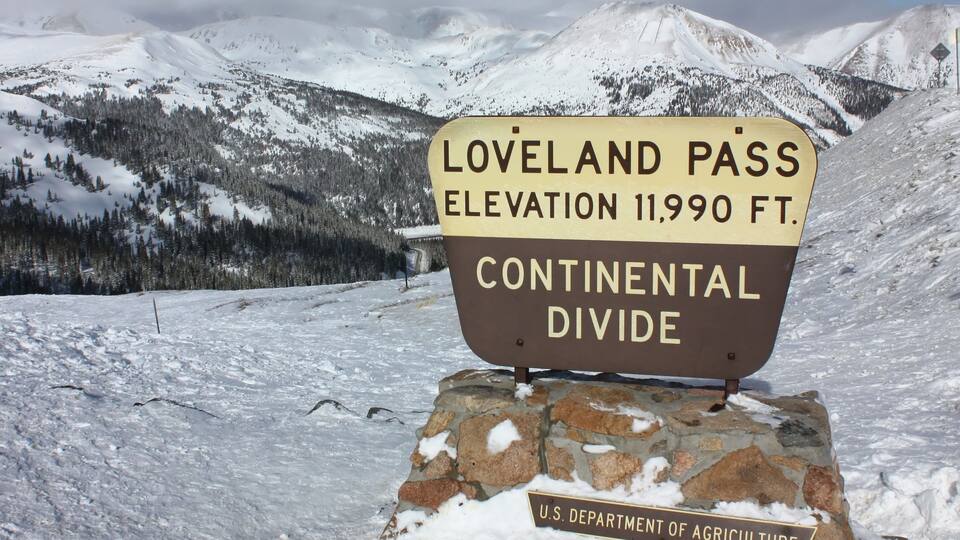 A sign indicating the elevation of Loveland Pass at 11,990 feet and its location on the Continental Divide, with a backdrop of snow-covered mountains and trees.