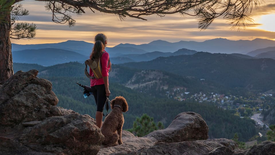 A hispanic woman is hiking with a dog, at sunset, in the Rocky Mountains near Denver, Colorado, USA