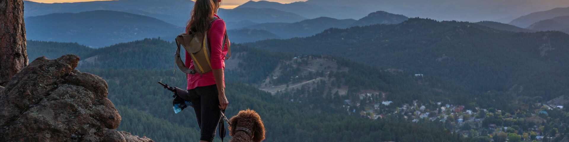 Uma pessoa com um cachorro em um afloramento rochoso, com vista para um vale e montanhas ao longe.