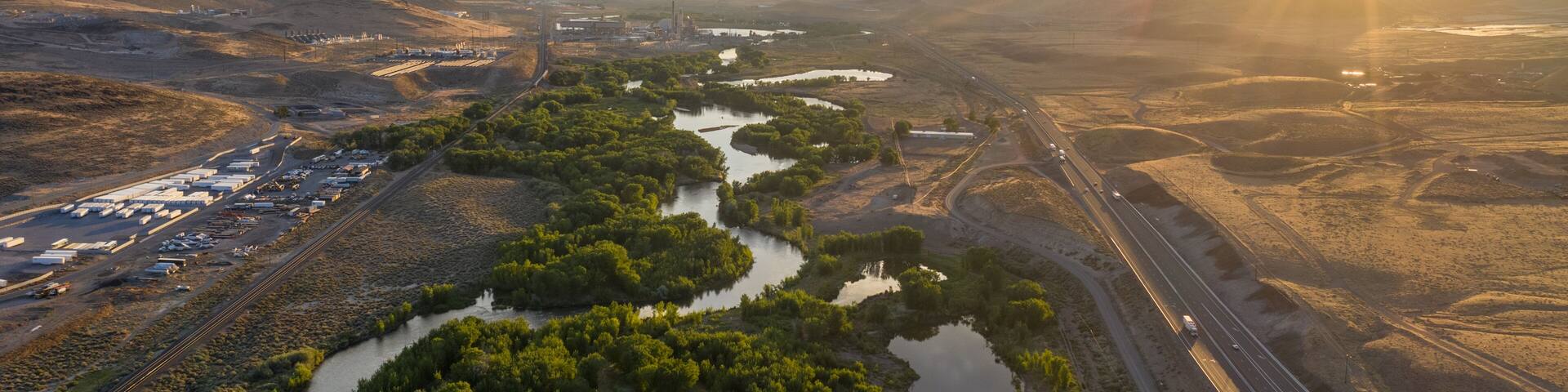A twisting turning tree lined river through a desert mountain range, a railroad and a major highway, and the sun setting over the horizon