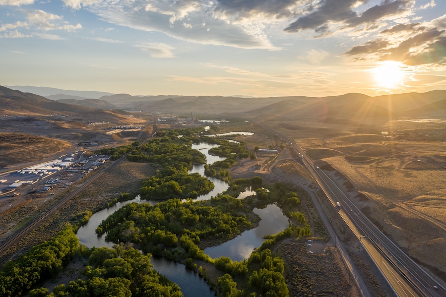 A twisting turning tree lined river through a desert mountain range, a railroad and a major highway, and the sun setting over the horizon