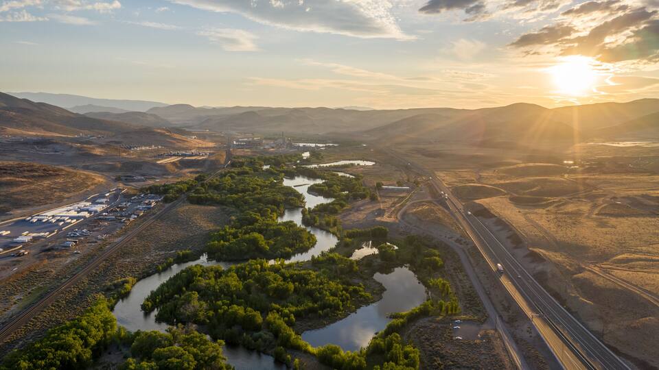 A twisting turning tree lined river through a desert mountain range, a railroad and a major highway, and the sun setting over the horizon