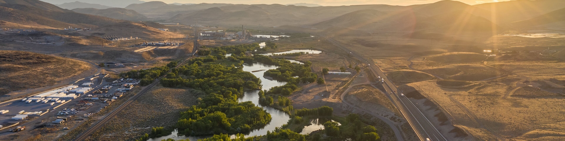 A twisting turning tree lined river through a desert mountain range, a railroad and a major highway, and the sun setting over the horizon