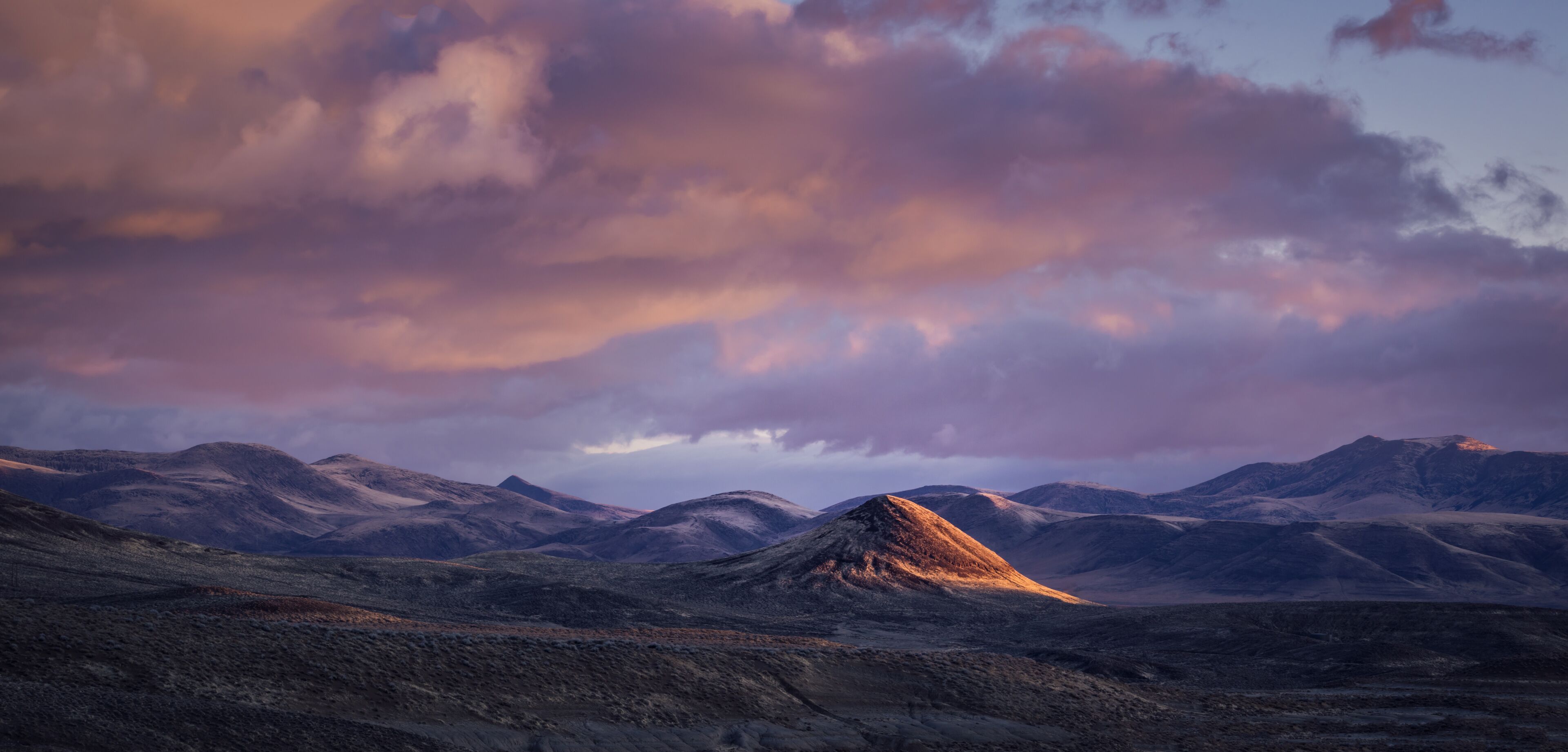 A mountainous landscape with a prominent peak in the center, surrounded by rolling hills and a dramatic sky with clouds.