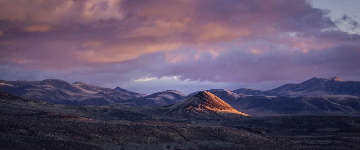 A mountainous landscape with a prominent peak in the center, surrounded by rolling hills and a dramatic sky with clouds.
