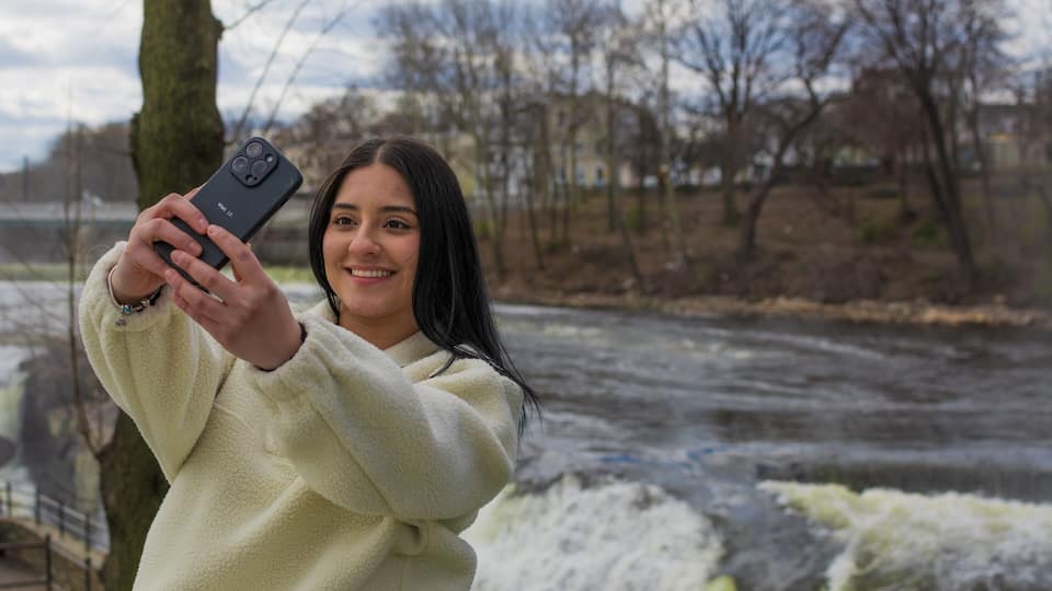 A person taking a selfie with a smartphone in a natural setting with a river and trees in the background.
