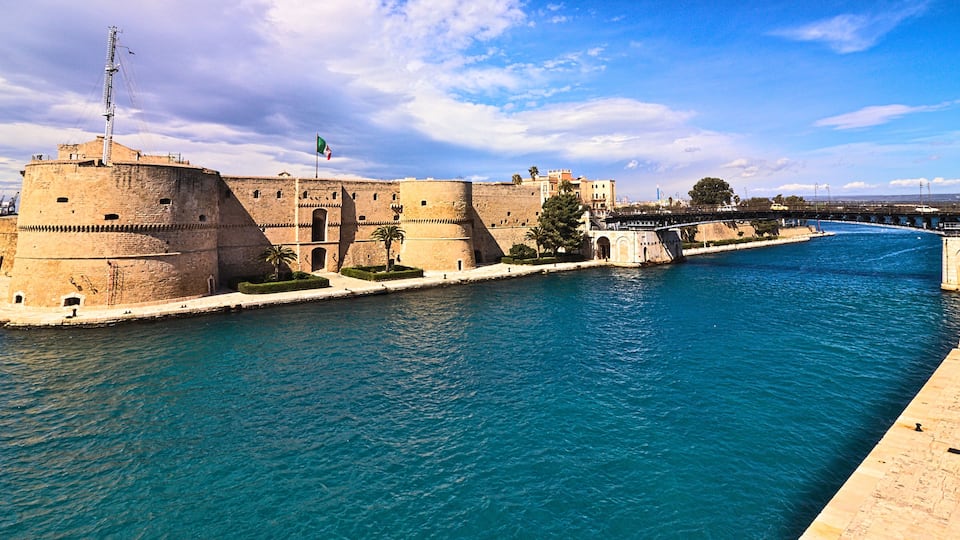 A historic fortification by a river with a bridge in the background.
