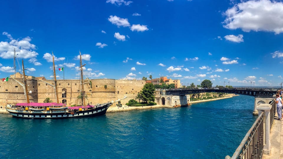A historic fort, a large sailing ship docked at the harbor, and a bridge connecting two land masses.