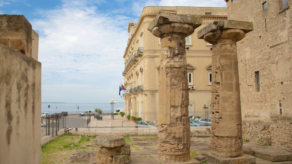 Ancient stone columns, a building with balconies, and a view of the sea.