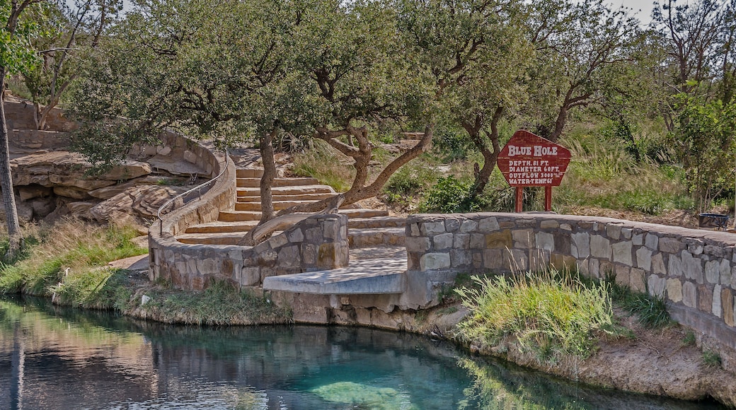 A stone bridge over a clear blue water body, surrounded by trees and a sign indicating 'Blue Hole' with depth and outflow details.